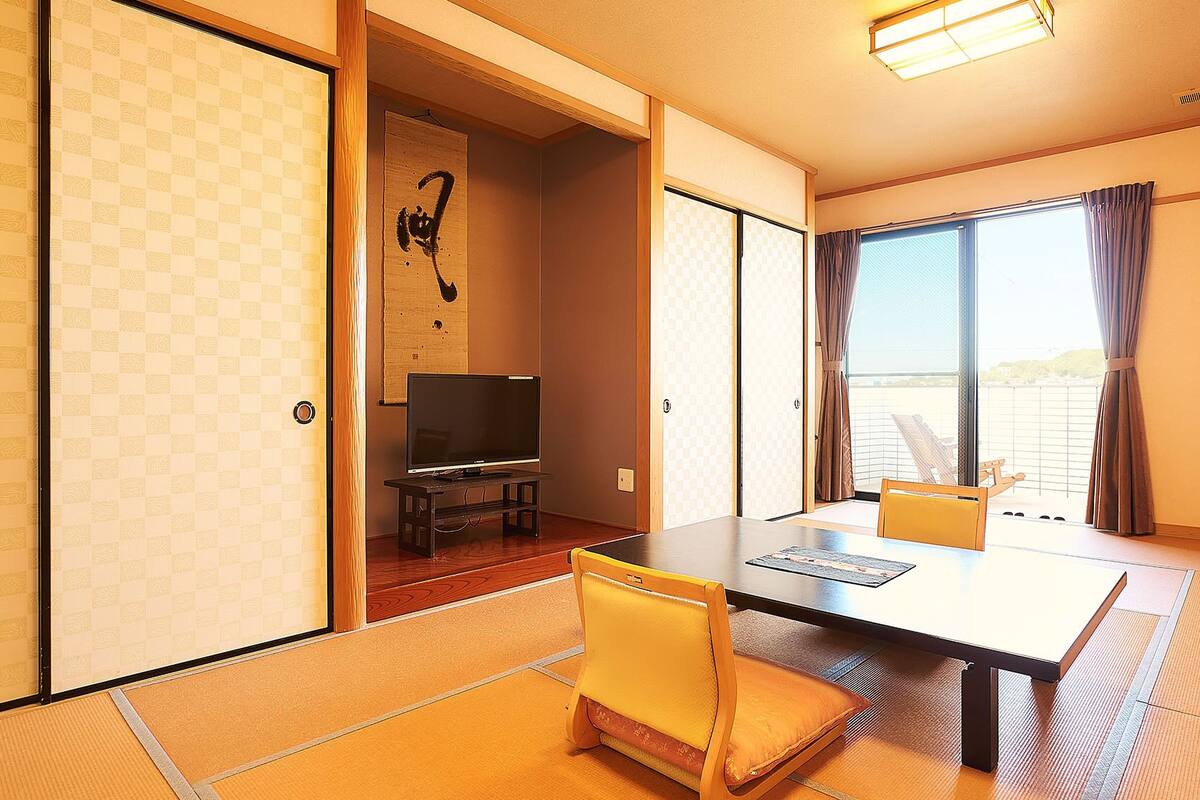 Ein traditioneller japanischer Raum mit Tatami-Böden, einem Fernseher auf einem niedrigen Standfuß und einer Schiebetür, die auf einen Balkon führt.