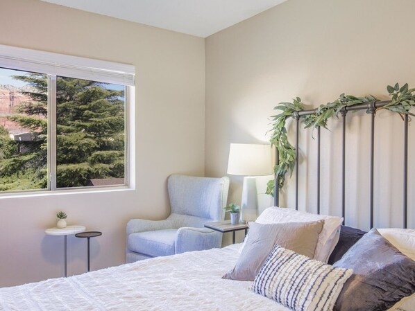 A bedroom with a bed, a chair, a small table, a lamp, and a window with a view of trees. - Stylish Sedona Condo | Red Rock Views, Modern Comfort & Quiet Location (Sedona)