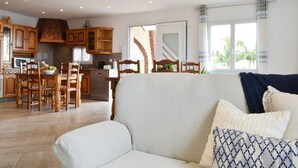 Amplia sala de estar con sofá y sillones, una mesa de comedor con sillas, armarios de madera y una zona de cocina con lavavajillas y microondas.