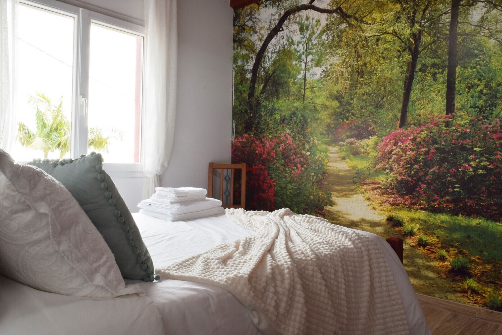 Una camera da letto con un grande murale raffigurante una foresta, un letto rifatto con lenzuola bianche e una finestra con tende.