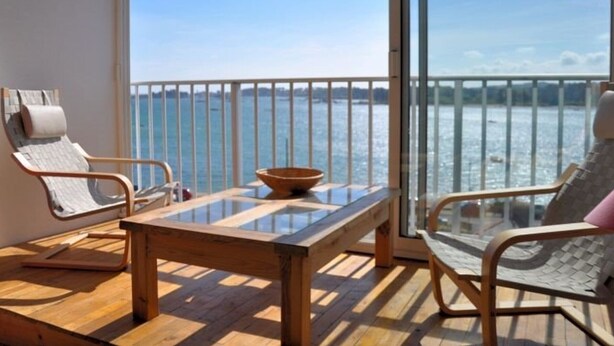 A balcony with a wooden table, two wicker chairs, and a glass balustrade overlooking a body of water.