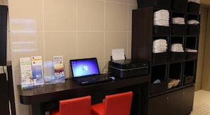 Een receptiebalie met een laptop, brochures en een printer. Er staan rode stoelen en een plank met opgevouwen handdoeken.
