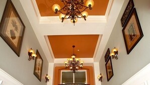 A hallway with a high ceiling, a chandelier, and a balcony. There are two wooden tables with chairs and potted plants. - Premium Lake View Condo [cdc Compliant] 1007 (Orlando)