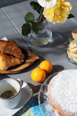 Uma mesa com croissants, laranjas, uma xícara de café, uma sobremesa com açúcar de confeiteiro e um vaso com flores.
