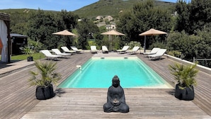 Una piscina rectangular con cubierta de madera, rodeada de sillas de descanso y sombrillas, y una estatua de Buda en el centro.