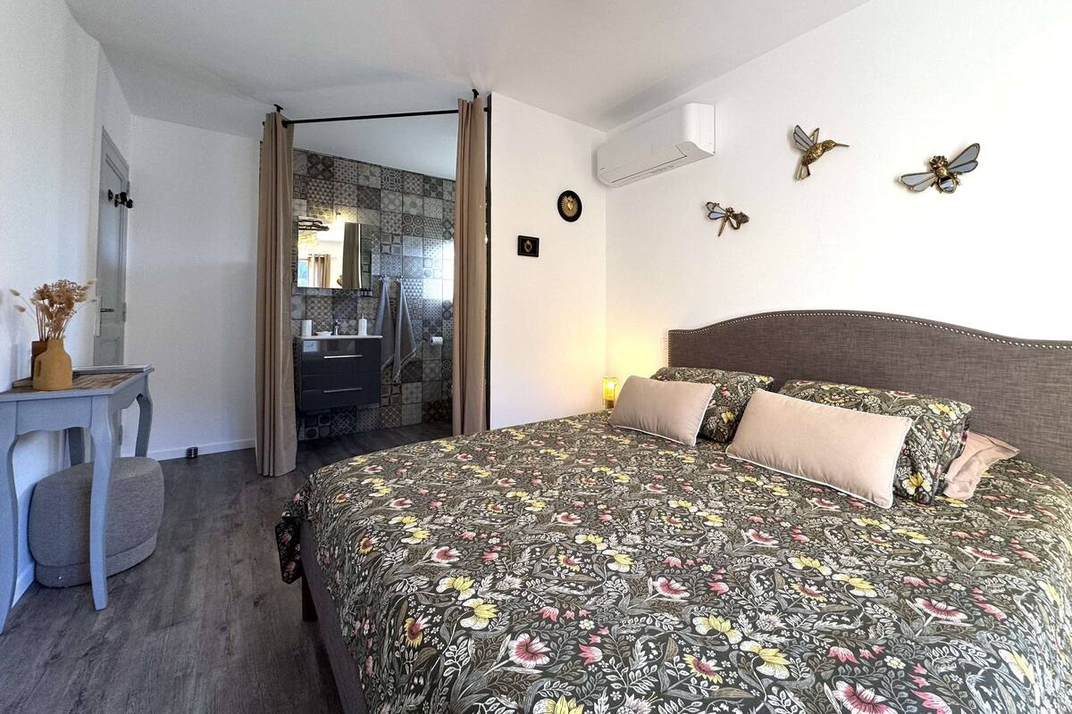 Une chambre à coucher avec une couvre-lit à motifs floraux, une table de chevet ornée d’un vase et une horloge fixée au mur.