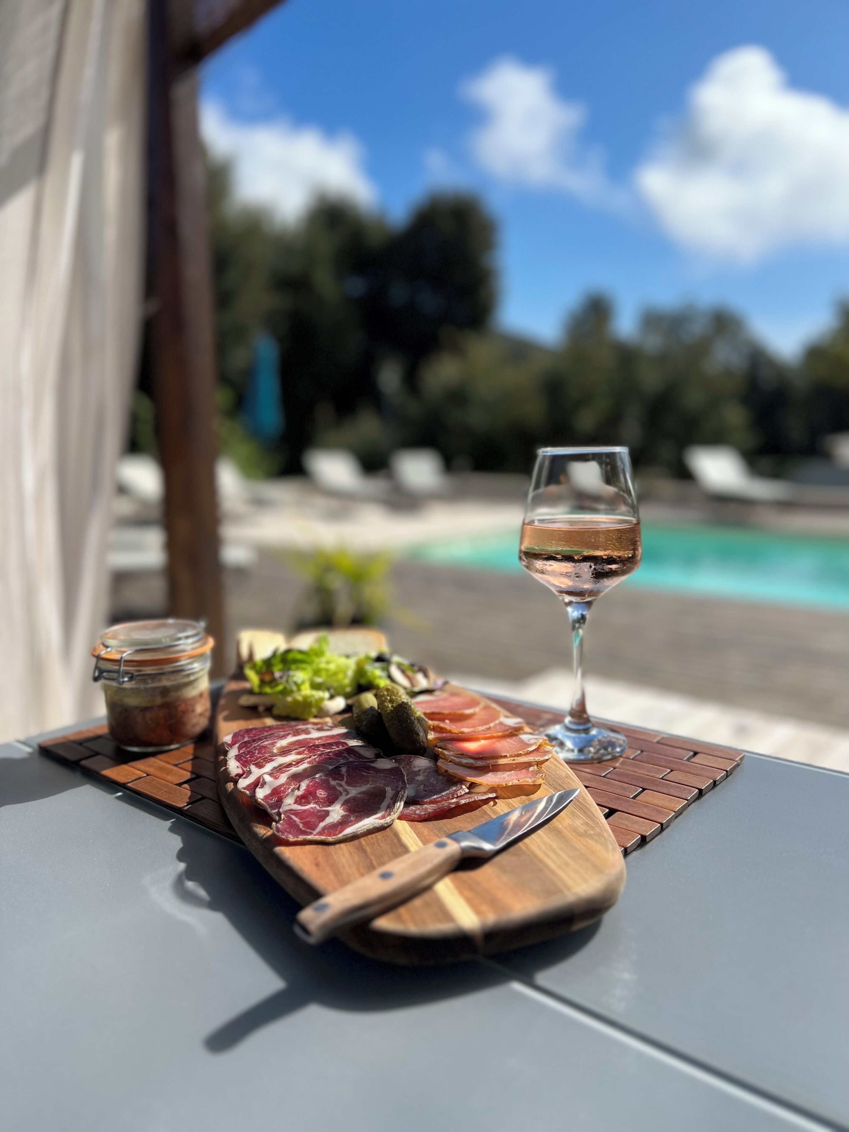 Una mesa con una tabla de embutidos, una copa de vino y un tarro de pepinillos.