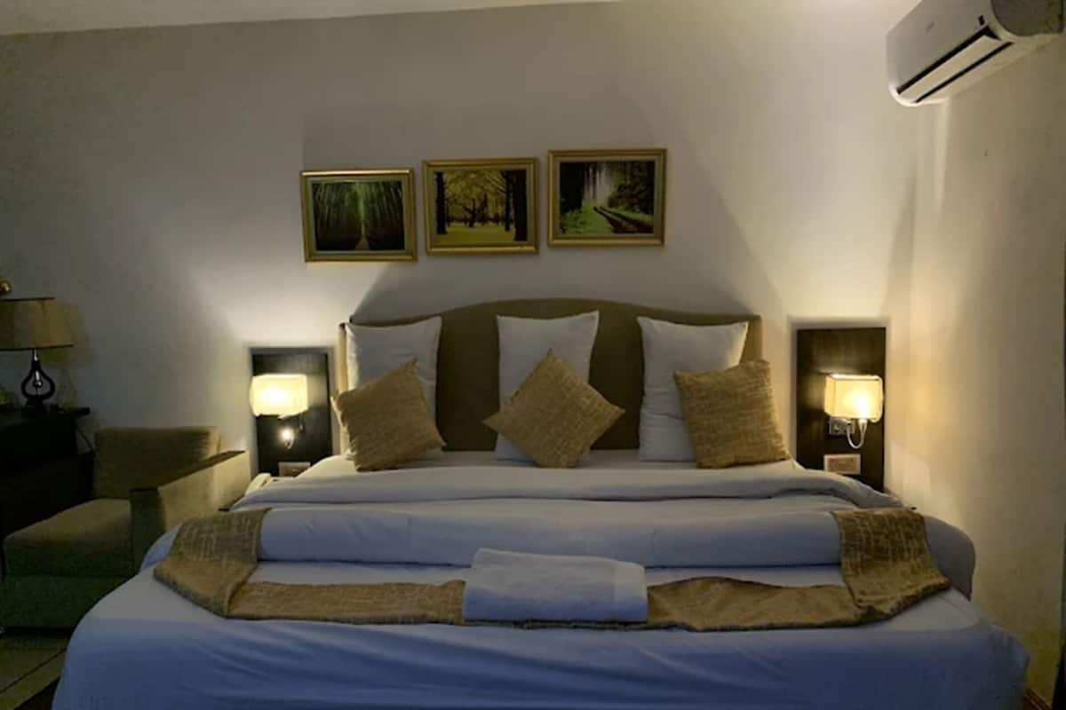 A neatly made bed with white linens and a beige throw blanket, flanked by two wall-mounted lamps and three framed pictures above.
