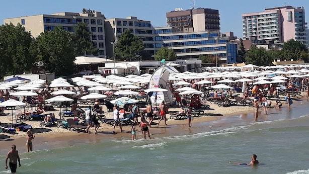 A beach with umbrellas and sun loungers, a few people in the water, and buildings in the background. - Krystal Park studio in Sunny Beach (Sonnenstrand)