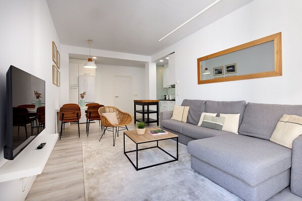 A modern living room with a grey sofa, a coffee table, a TV, and a dining area with chairs and a table.