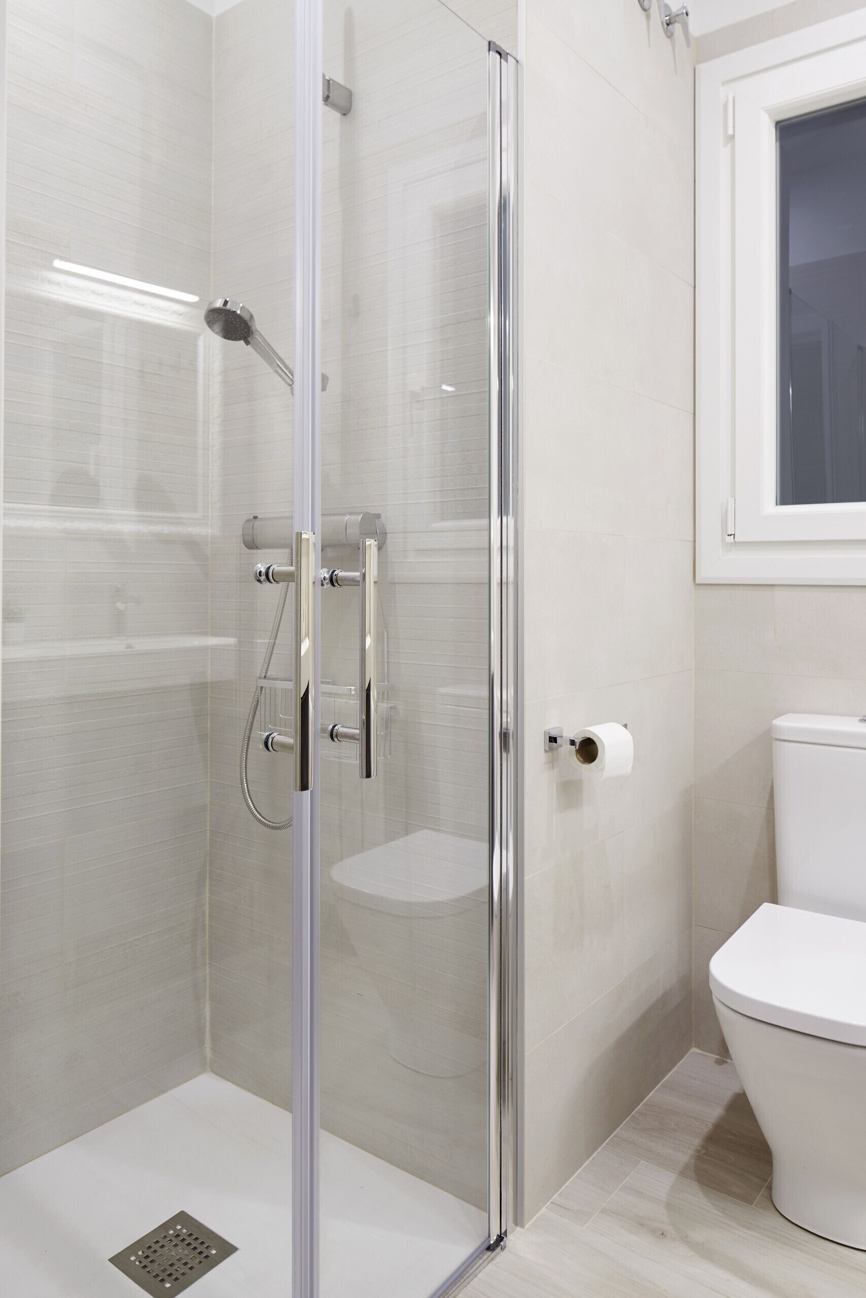 A modern bathroom with a glass-enclosed shower, a toilet, and a window.