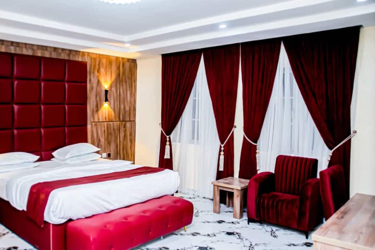 A hotel room with a large bed, red upholstered headboard, two bedside lamps, a red chair, and a small wooden table.