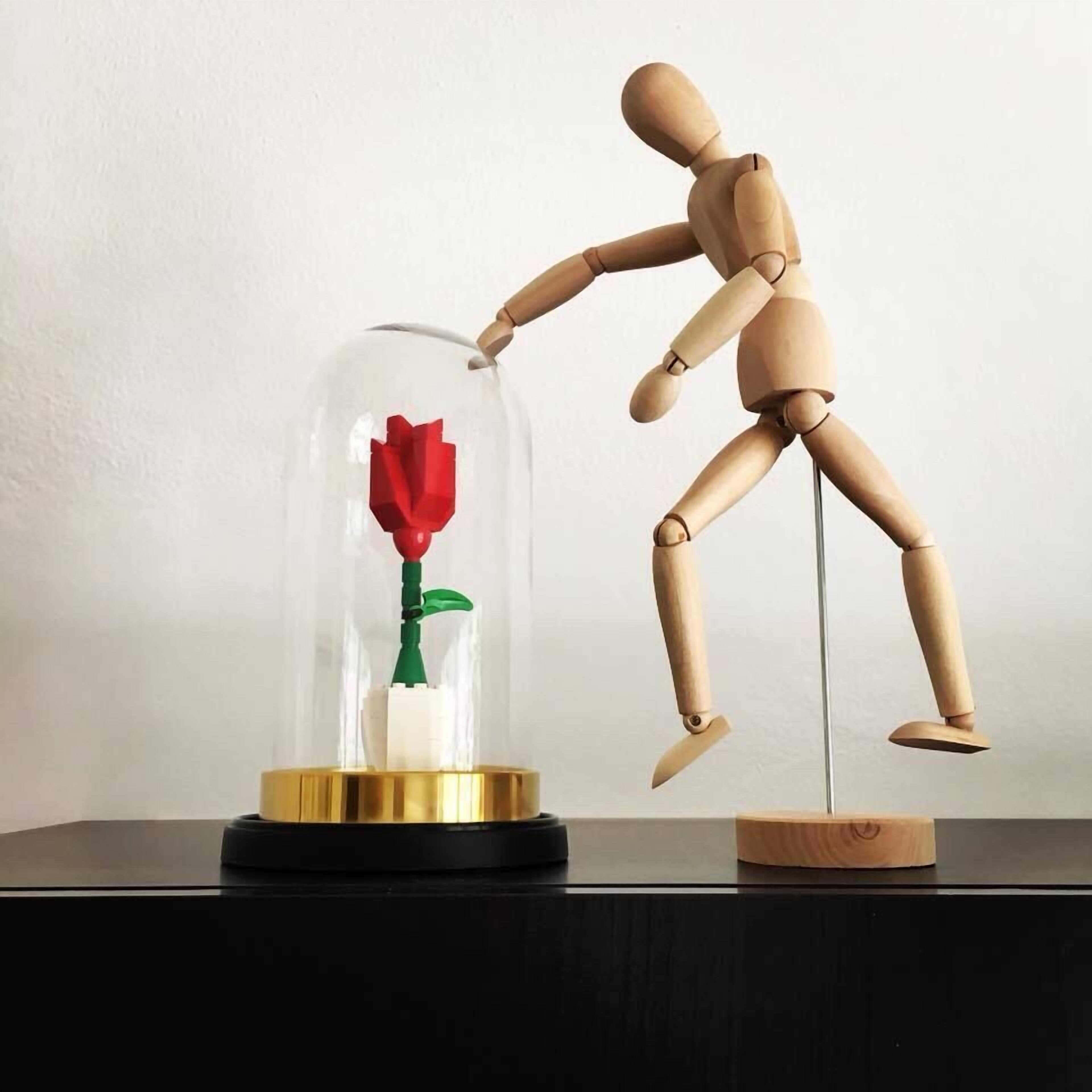 Un mannequin en bois appuyé contre une cloche de verre contenant une rose rouge.