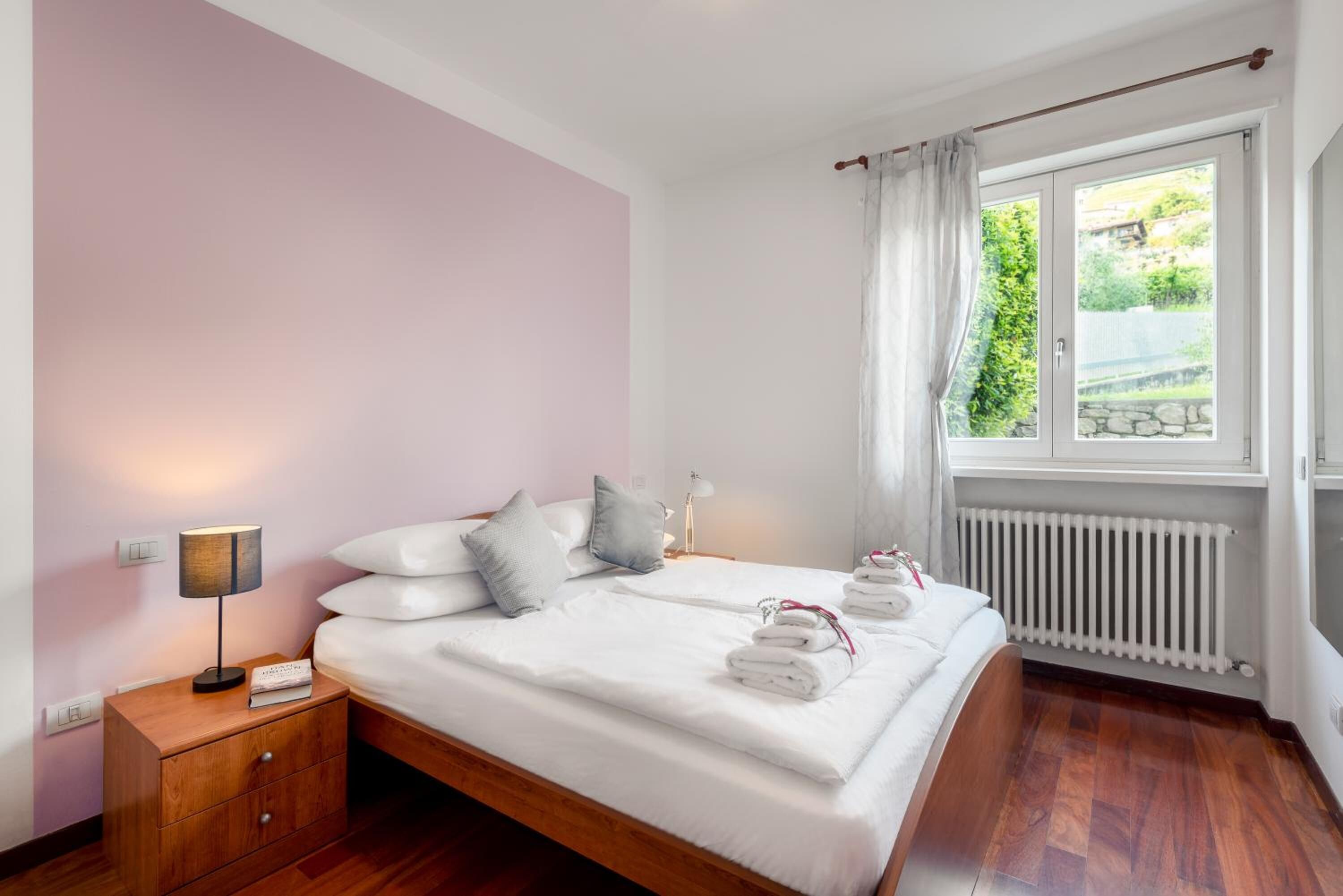 Une chambre à coucher avec un lit en bois, du linge de lit blanc, une table de chevet avec une lampe, et une vue sur la verdure par la fenêtre.