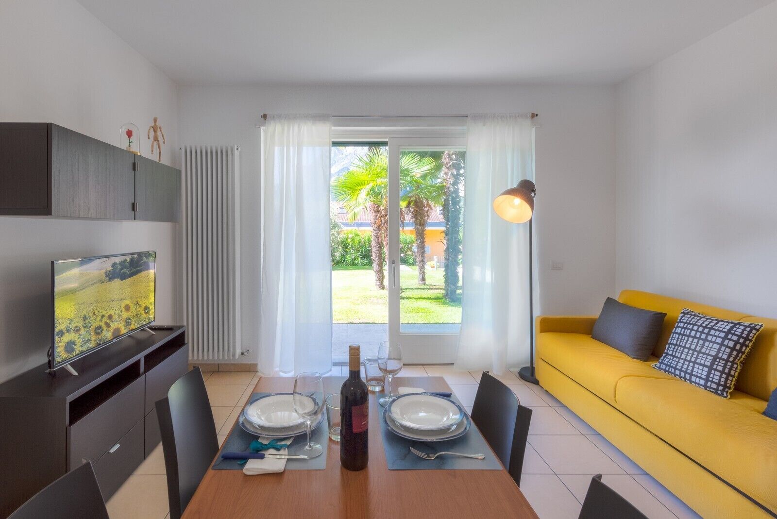 A modern dining area with a table set for two, a sofa, a TV, and a view of a garden through a glass door.