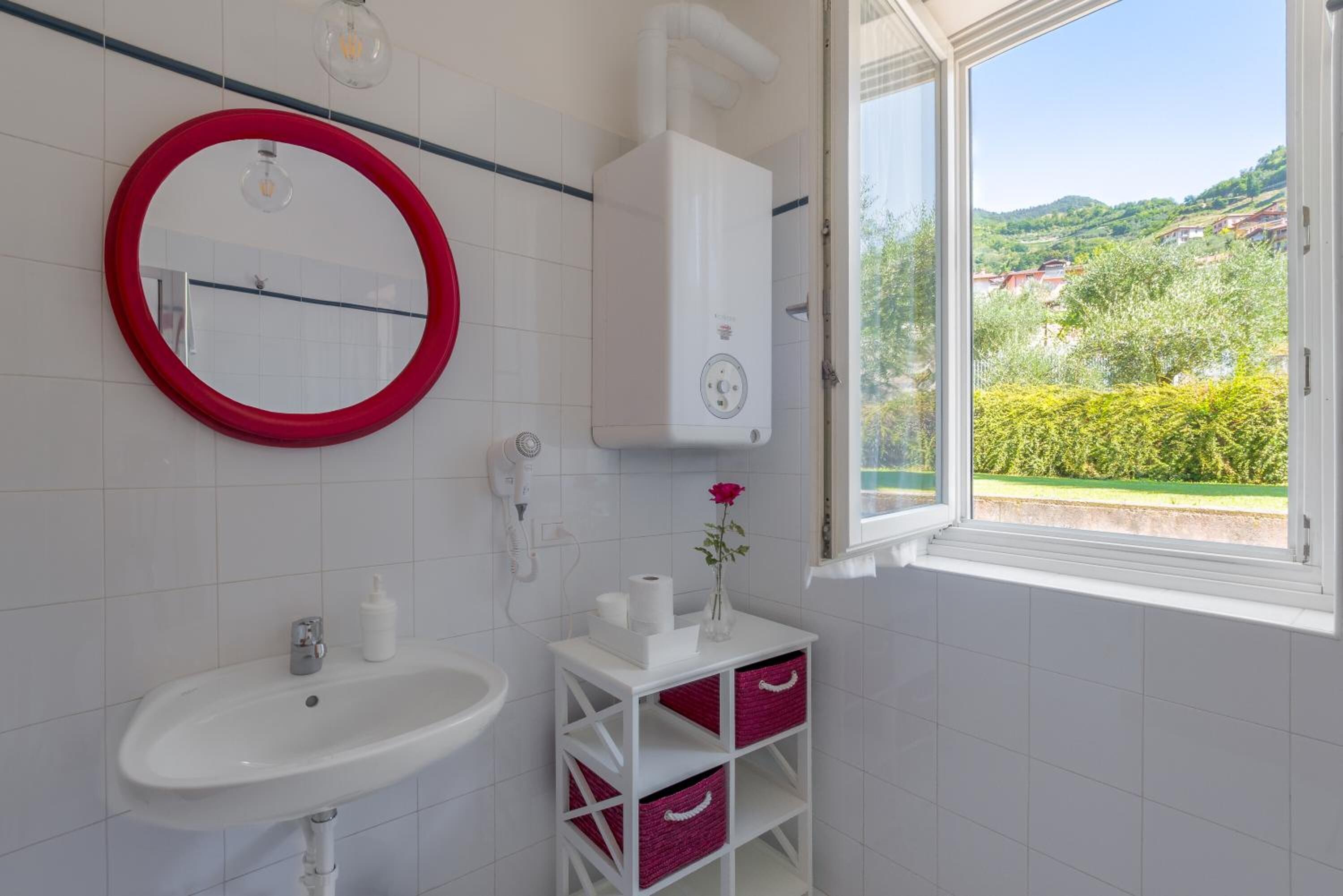 A bathroom with a white sink, a red-framed mirror, a white heater, and a window with a view of greenery and houses.