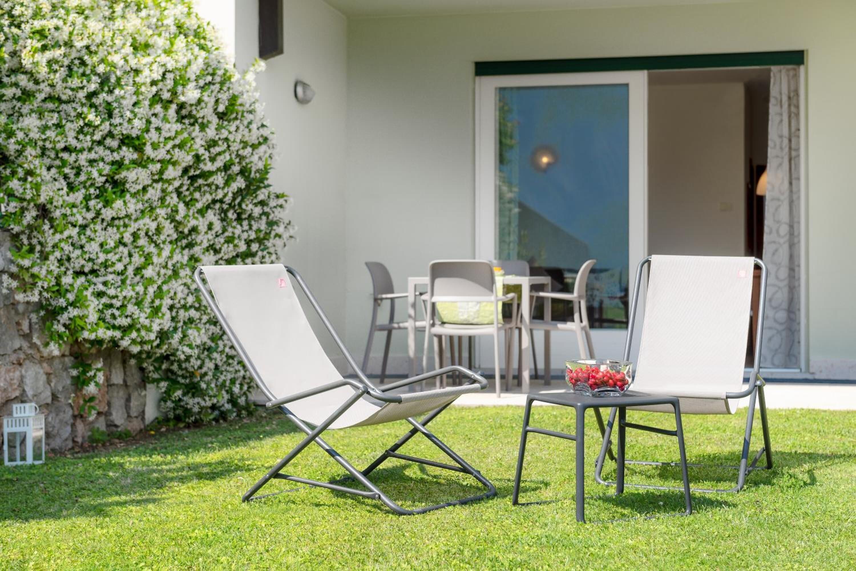 An outdoor seating area with two chairs and a table, a stone wall with white flowers, and a building entrance with a glass door.