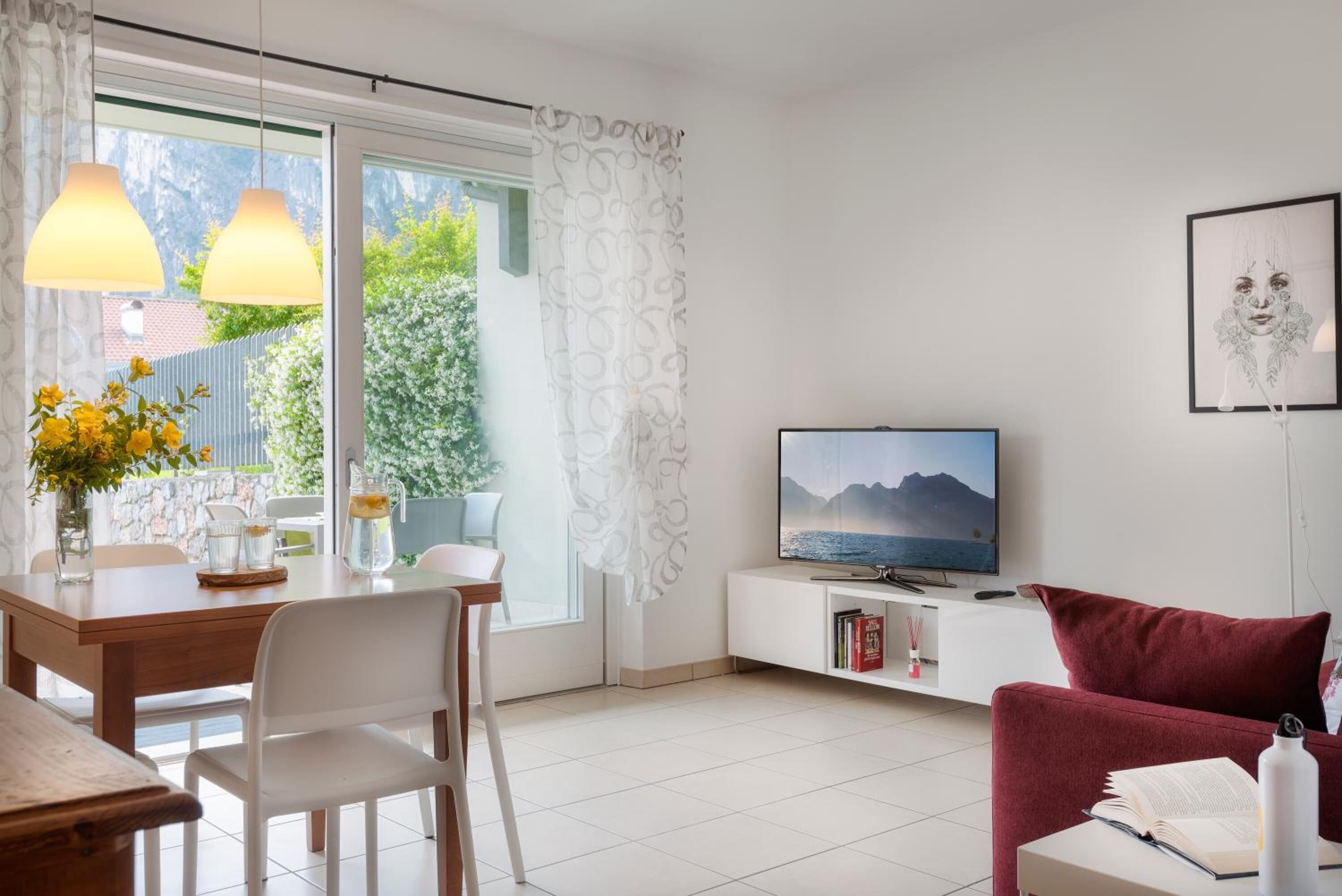 A modern living room with a TV, a red sofa, a dining table with chairs, and a view of greenery outside.