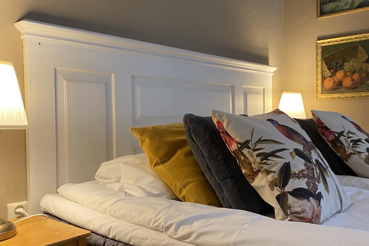 Ein ordentlich bezogenes Bett mit weißer Bettwäsche, ein Nachttisch aus Holz mit einer Lampe und ein an der Wand hängendes Bild.