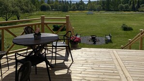 Una terraza de madera con mesa y sillas, un gran paraguas y vistas a un campo cubierto de césped y árboles.