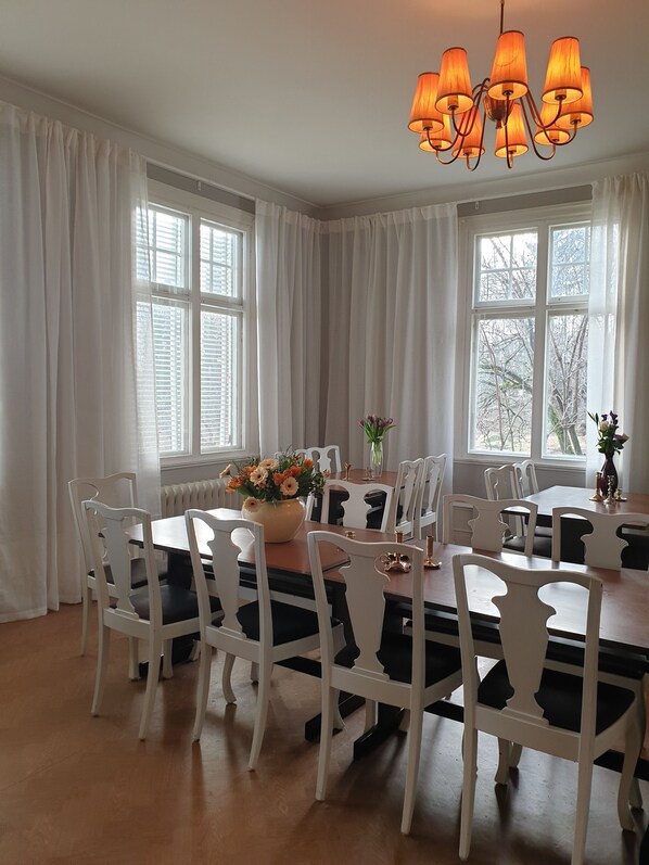 A dining room with a wooden table and white chairs, large windows with white curtains, and a chandelier.
