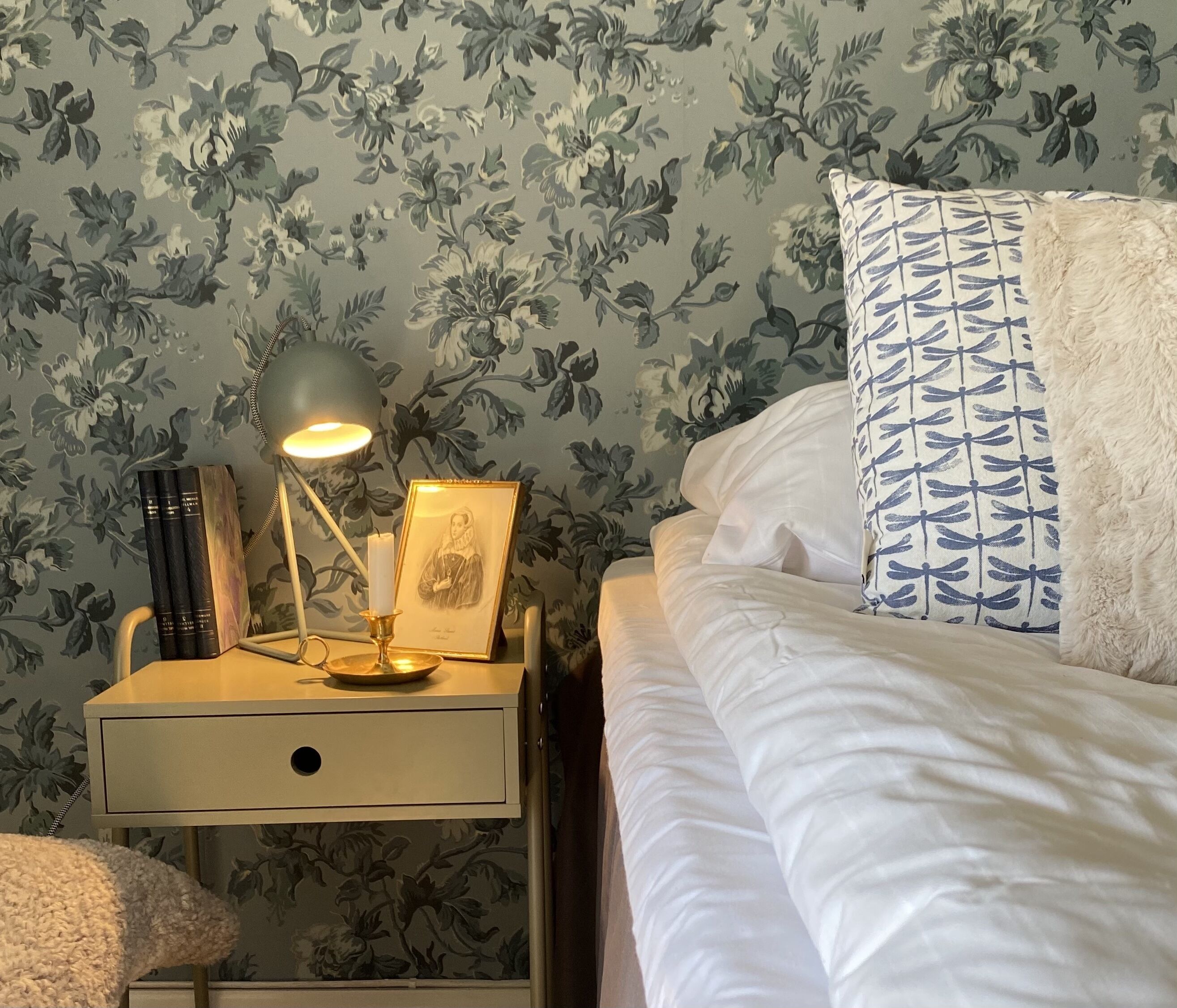 A neatly made bed with white bedding, a bedside lamp, a framed picture, and a stack of books on a nightstand.