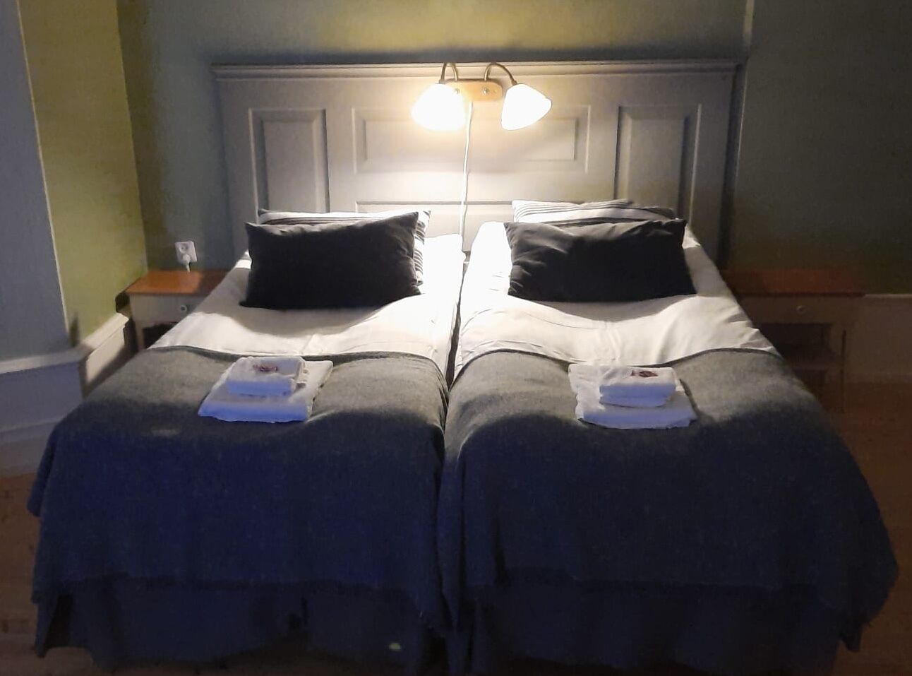 Una habitación con dos camas individuales, cada una con una almohada y una toalla doblada. Hay una mesita de noche con una lámpara entre las camas.