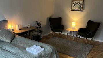 A hotel room with a bed, two chairs, a small table, a desk, and a plant.