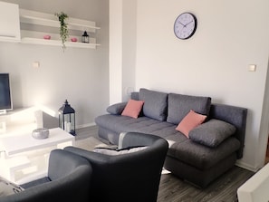 A modern living room with a grey sofa, black armchairs, a white coffee table, a TV, and a wall clock.
