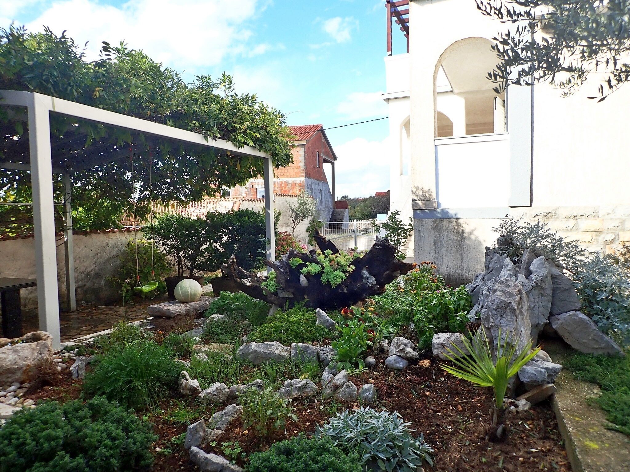 Un giardino con diverse piante e rocce, un pergolato ricoperto di piante rampicanti e un edificio bianco con tetto rosso sullo sfondo.