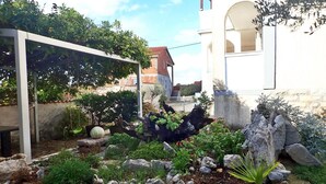 En trädgård med olika växter och stenar, en pergola med klätterväxter och en vit byggnad med ett rött tak i bakgrunden.
