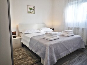 A neatly made bed with white linens and a grey quilt, a bedside table with a lamp, and a small framed picture on the wall.