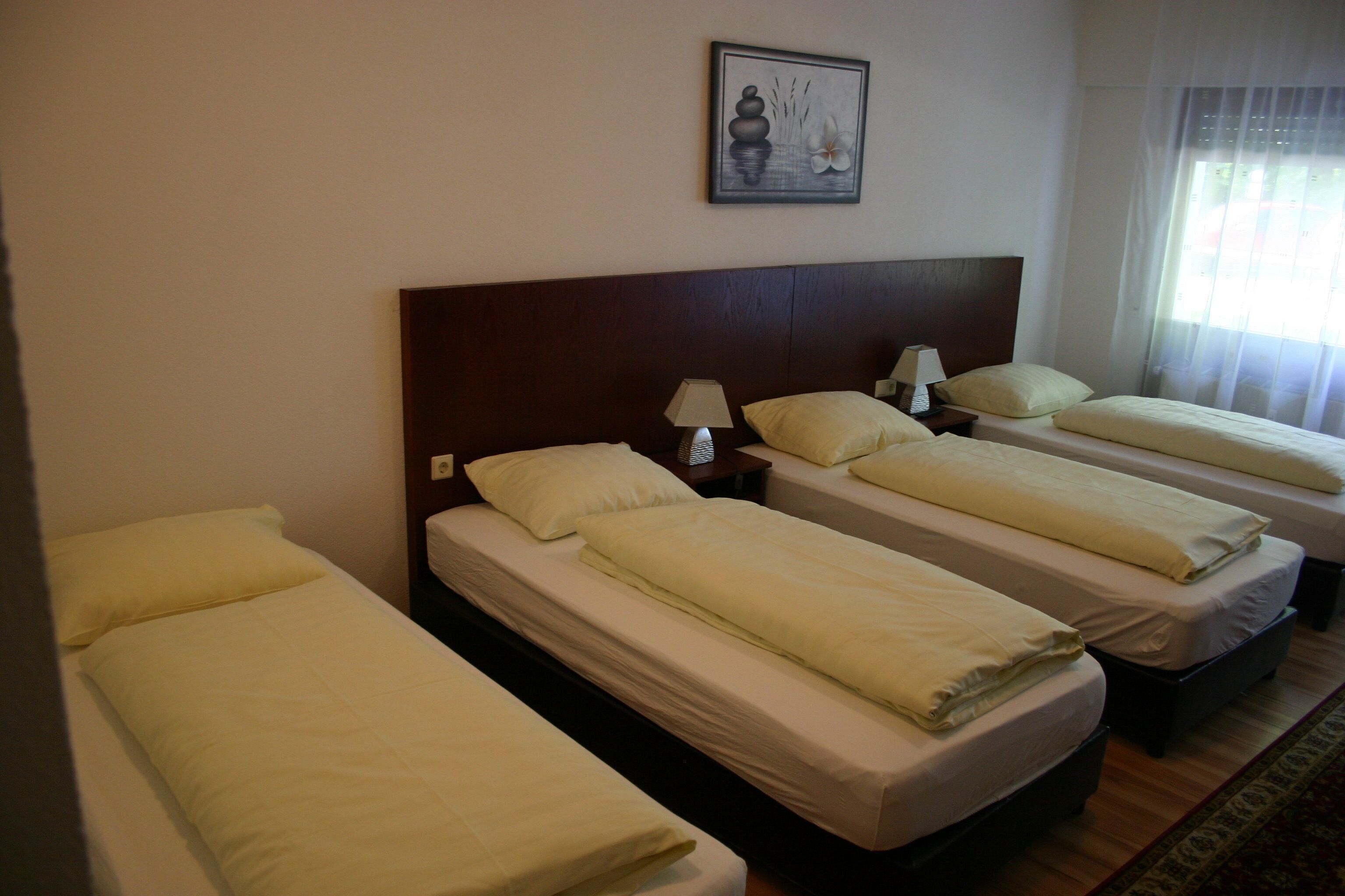A hotel room with four single beds, each with a wooden headboard, white bedding, and a bedside table with a lamp.
