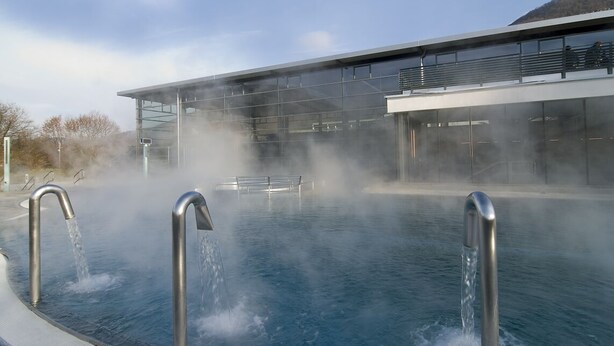 Un edificio moderno con fachada de vidrio y una piscina con vapor ascendiendo del agua.