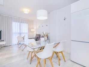 Ein moderner, minimalistischer Wohn- und Essbereich mit Glastisch, weißen Stühlen, einer Couch und einem weißen Kühlschrank.