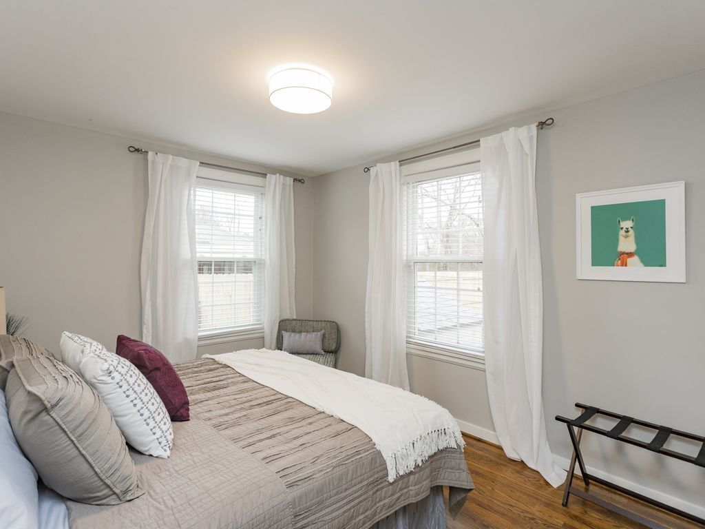 Un dormitorio con cama, dos ventanas con cortinas blancas, una silla, un banco y un cuadro en la pared.