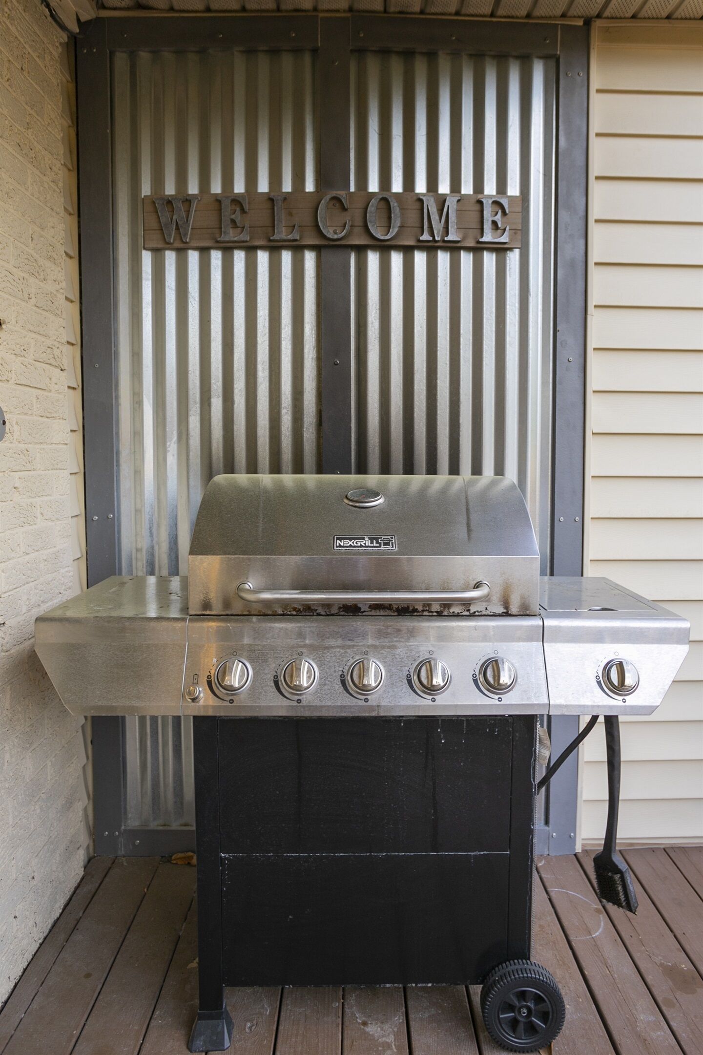 Una parrilla de acero inoxidable con cuatro quemadores y un panel de control, ubicada en una terraza de madera con un letrero que dice 'WELCOME'.
