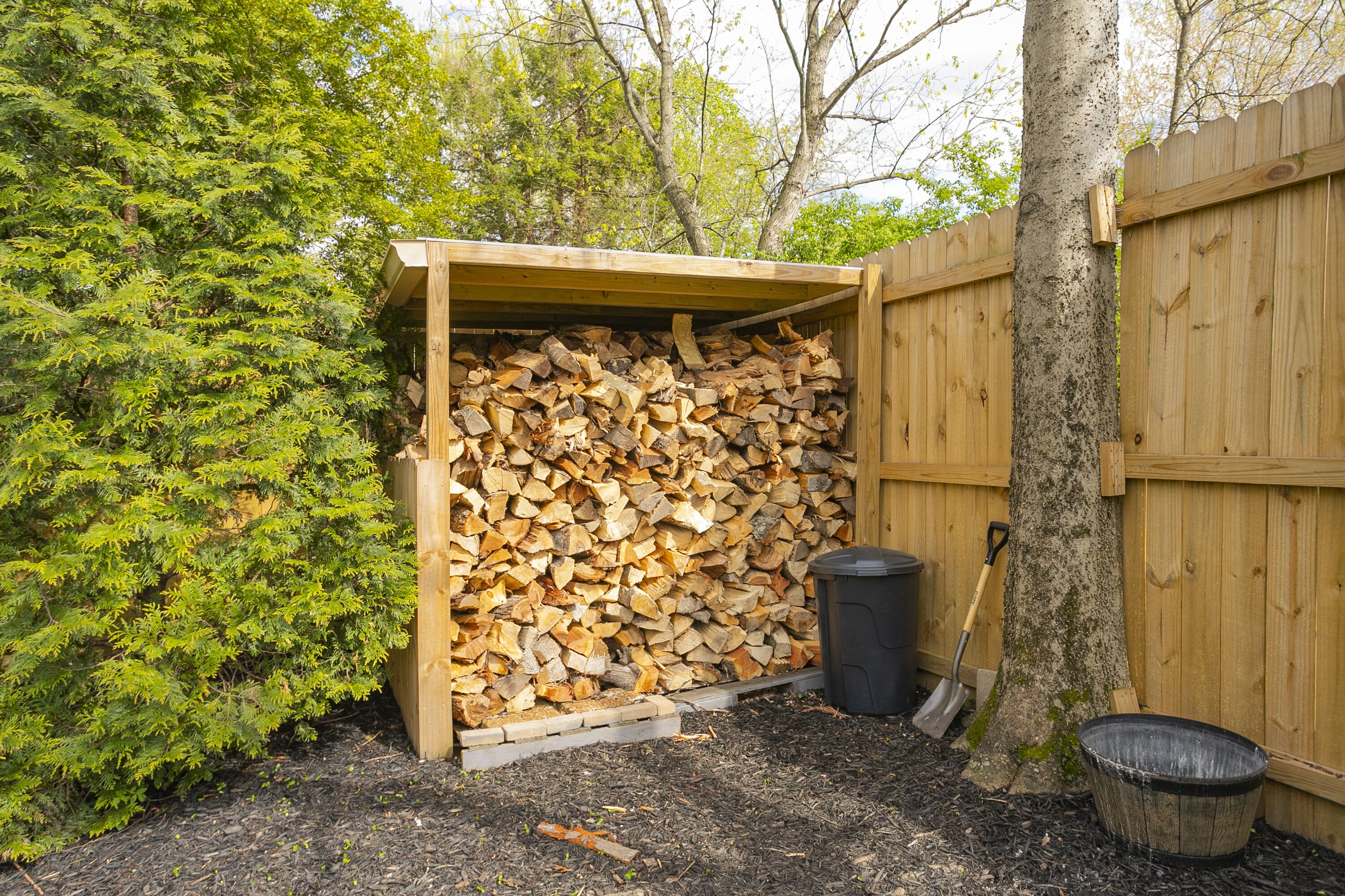 Ein hölzernes Gartenhaus mit einem Holzstoß darin, eine Schaufel, die an der Hauswand lehnt, und einer schwarzen Mülltonne daneben.