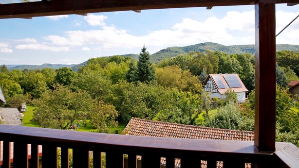 A wooden balcony with a view of a house with solar panels, surrounded by lush greenery and hills. - Apartment 1 - Apartment am-edersee (Edertal)