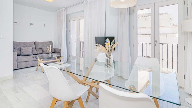 A modern living room with a glass dining table, white chairs, a grey sofa, and a TV.