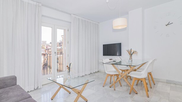 A modern living room with a glass dining table, white chairs, a grey sofa, a flat-screen TV, and a large window with curtains. - Spacious and modern Apartment with free parking in the heart of Granada 10 (Granada)