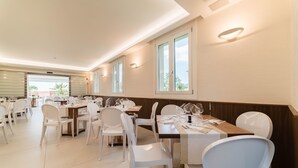 Un vaste espace dédié à la salle à manger, avec de nombreuses tables et des chaises blanches, de grandes fenêtres et un éclairage encastré au plafond.