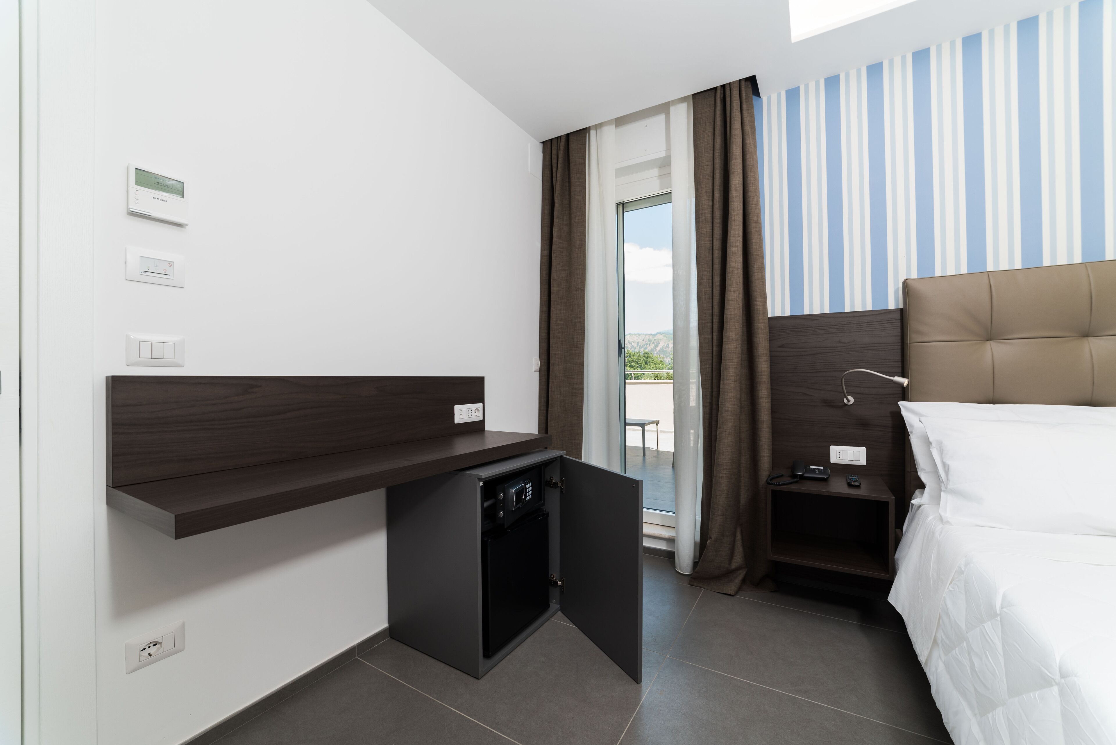 A hotel room with a bed, a desk with a small refrigerator, a nightstand, and a view of the outdoors.