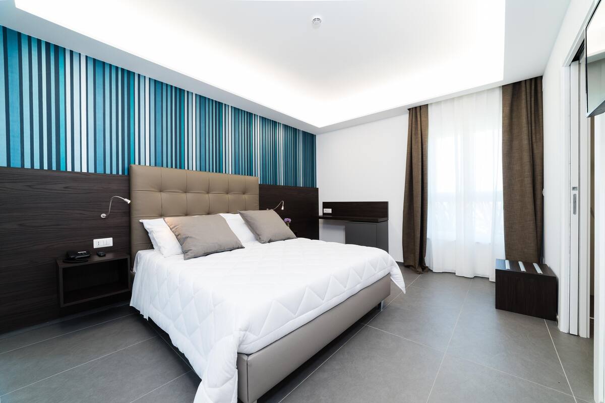 A modern hotel room with a large bed, a nightstand, a desk, and a wardrobe.