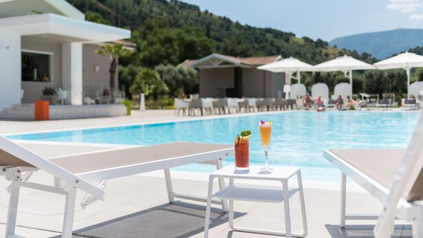 Un espace aménagé au bord de la piscine, avec des chaises longues et des parasols, une table avec des boissons, et une vue sur une montagne et la piscine.