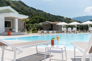 A poolside area with lounge chairs and umbrellas, a table with drinks, and a view of a mountain and pool. - Likò Resort (Nocera Inferiore)