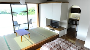 A compact room with a tatami mat floor, a bed with a plaid blanket, a small table, a TV, and a kitchenette with a microwave. - Twin House Umigukuru (Nakijin)