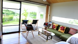 A modern living room with a large sliding glass door leading to a balcony, a green sofa with colorful cushions, and two folding chairs. - Twin House Umigukuru (Nakijin)