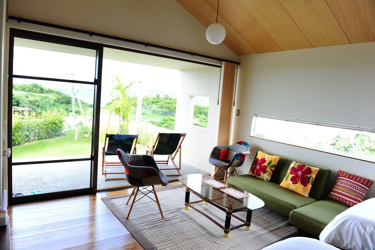 A modern living room with a large sliding glass door leading to a balcony, a green sofa with colorful cushions, and two folding chairs.