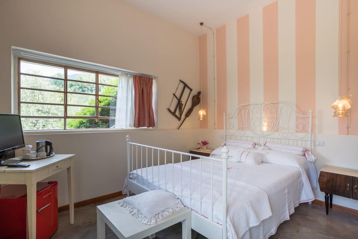 A bedroom with a bed, a desk with a computer, a window with curtains, and a wall with striped wallpaper.
